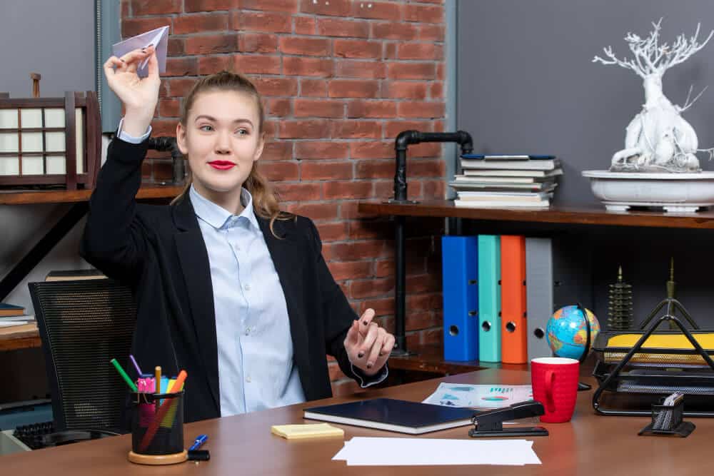 Top view of young smiling female office worker sitting at her desk and playing paper plane